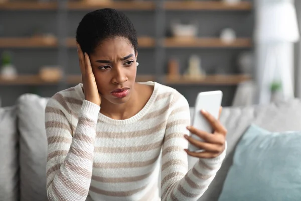 Orolig svart kvinna tittar på Smartphone när du sitter på soffan hemma — Stockfoto