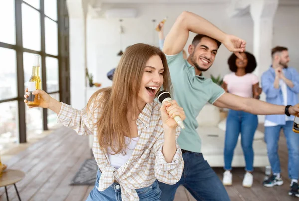 Jovem caucasiana alegre com garrafa de cerveja cantando karaoke em festa estudantil com seus amigos em casa — Fotografia de Stock