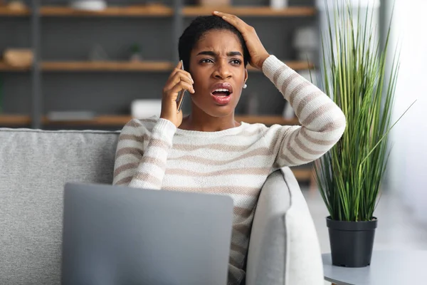 Problema con Internet. Mujer negra confusa hablando en el teléfono celular y usando el ordenador portátil —  Fotos de Stock