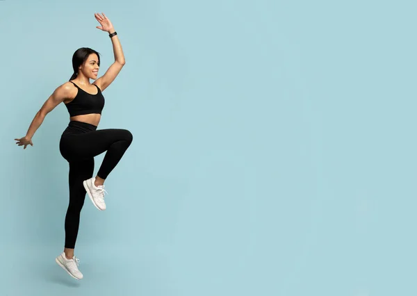 Concepto fitness. Ajuste joven afroamericana mujer saltando, levantando la pierna hacia arriba, fondo azul con espacio vacío —  Fotos de Stock