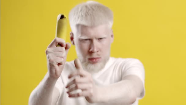Парень-альбинос позирует с бананом, делая вид, что стреляет из ружья, жёлтый фон — стоковое видео