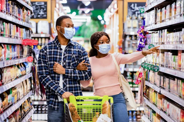 Νεαρό μαύρο ζευγάρι με μάσκες προσώπου περπατώντας μέσα από σειρές σούπερ μάρκετ με καλάθι αγορών, δείχνοντας στα ράφια, επιλέγοντας τα τρόφιμα — Φωτογραφία Αρχείου