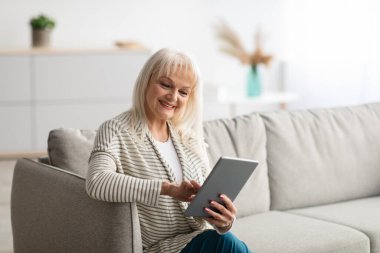 Kanepeye oturmuş tablet kullanan gülümseyen olgun bir kadın.