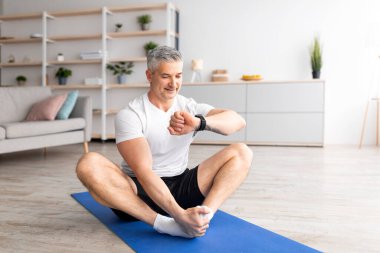 Eğitim zamanı. Olgun bir adam elindeki fitness izleyicisine bakıyor, evde spor paspası üzerinde egzersiz yapıyor.