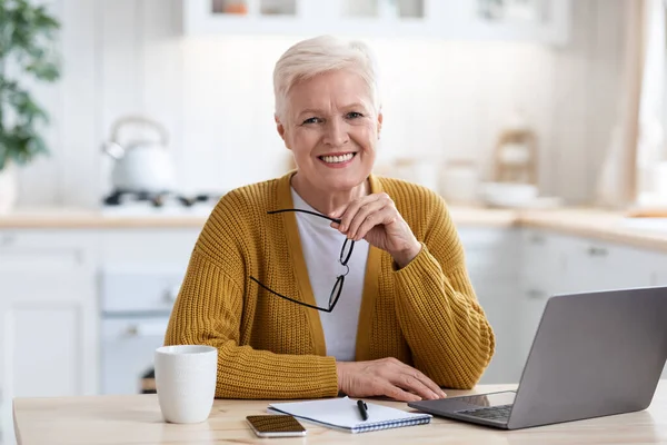 Fröhliche Seniorin sitzt vor Laptop, Kücheneinrichtung — Stockfoto