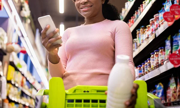 Närbild av svart dam kontroll inköpslista i mobilappen, sätta flaska mjölk i vagnen på köpcentret, selektivt fokus — Stockfoto
