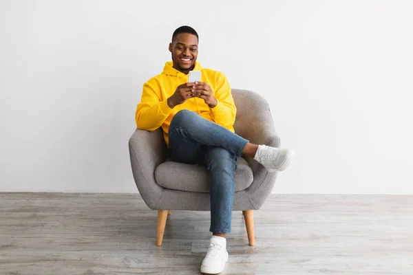 Africano chico usando smartphone sentado en silla sobre gris pared —  Fotos de Stock