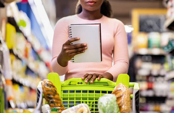 アフリカ系アメリカ人女性ショッピングカートスーパーマーケットでノートブックに食料品リストをチェックする製品のフル — ストック写真