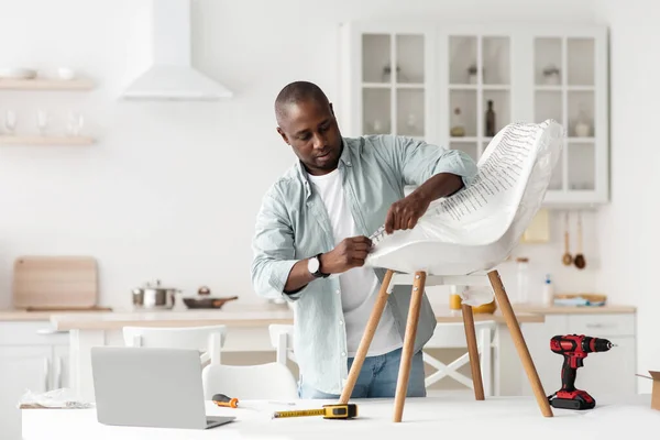 Hombre afroamericano recibiendo muebles nuevos, desempacando la silla y usando el ordenador portátil en el interior de la cocina, espacio de copia — Foto de Stock