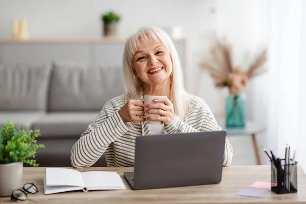 Volwassen vrouw zitten aan bureau drinken koffie werken op de computer — Stockfoto
