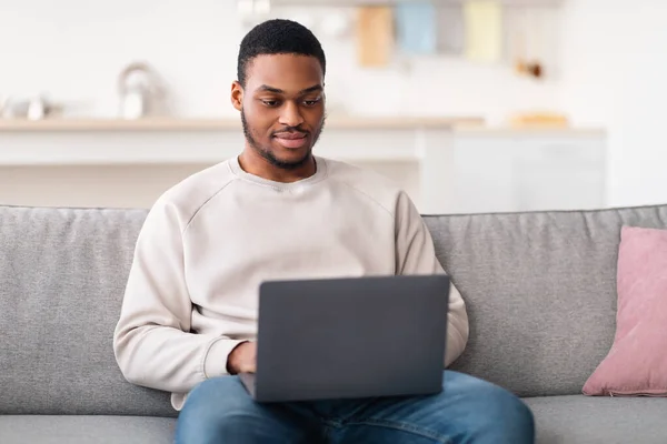 Sonriente chico negro usando el ordenador portátil, sentado en el sofá —  Fotos de Stock