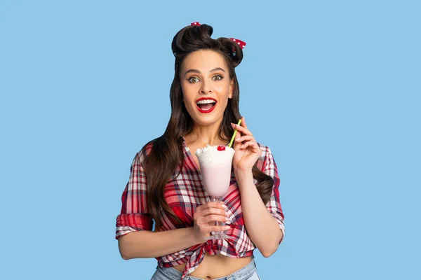 Porträt einer glamourösen Pinup-Frau im Retro-Stil, die sich glücklich fühlt, während sie Milchshake vor blauem Hintergrund trinkt — Stockfoto