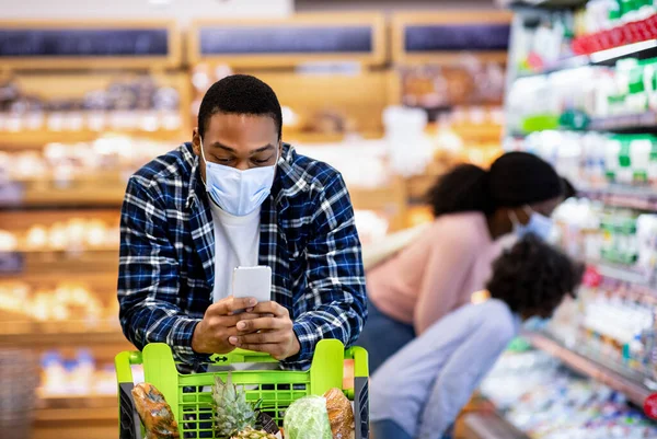 黒男で顔マスクチェック食料品リスト上のスマートフォン一方で彼の妻と娘選択製品にモール — ストック写真