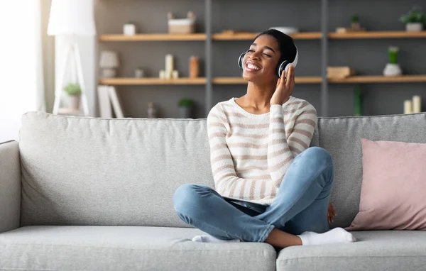 週末はリラックス。家庭でのワイヤレスヘッドフォンでオーディオブックを聞く幸せな黒人女性 — ストック写真