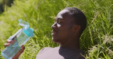 Su dengesi kavramı. Genç, susamış Afrikalı Amerikalı adam plastik şişeden taze su içiyor, çimlerin üzerinde yatıyor.