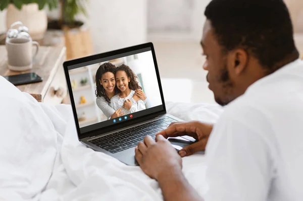 Ung svart man talar till fru och dotter på webbkamera, har online videokonferens på bärbar dator, liggande på sängen hemma — Stockfoto