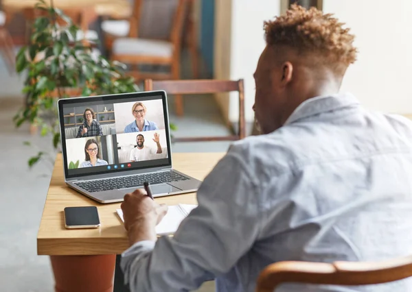 Black man having online work meeting with group of colleagues on laptop, taking notes at cafe — Stock Photo, Image