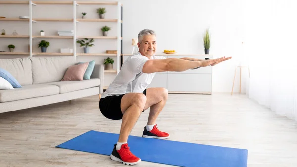 Home workout concept. Happy senior man in sportswear doing squats indoors in living room interior — стоковое фото