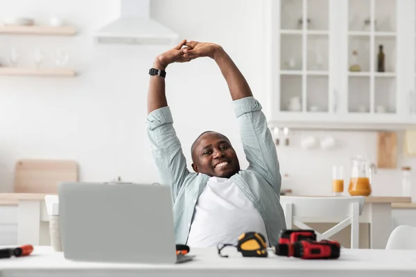 Gelukkig zwart man stretching handen tijdens het gebruik van laptop computer, zitten in de keuken met verschillende tools op tafel — Stockfoto