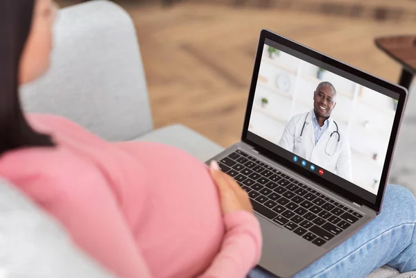 Online medical consultation. Expectant woman having video call with friendly black doctor on laptop from home