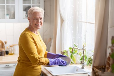 Mutfakta bulaşık yıkayan neşeli yaşlı bayan, lastik eldiven kullanıyor.