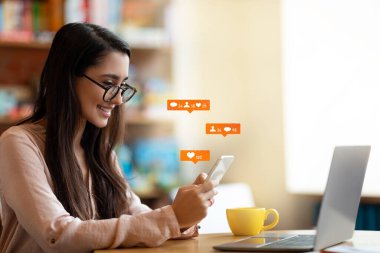 Sosyal medya bildirim simgeleriyle akıllı telefon kullanan mutlu Latin kadın blogcu, kafede oturuyor, boş alan.