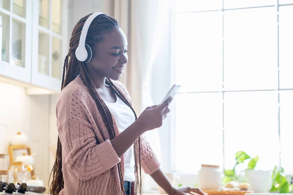 Alegre hembra negra en auriculares inalámbricos escuchando música en el teléfono inteligente en la cocina —  Fotos de Stock