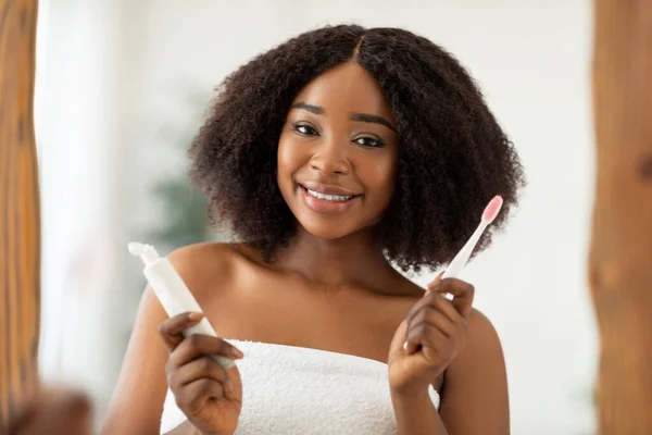 Cuidados dentários. Mulher negra jovem atraente com sorriso radiante segurando pasta de dentes e escova na frente do espelho em casa — Fotografia de Stock