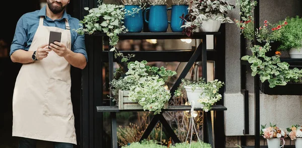 Young smiling bearded caucasian guy seller in apron typing on smartphone at front door of shop