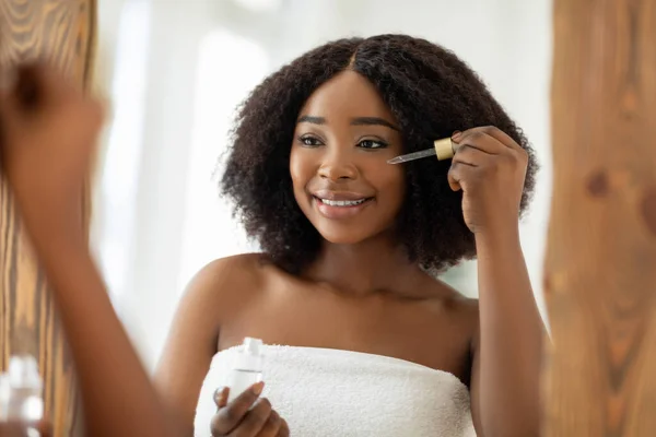 Kosmetik kecantikan untuk kulit muda. Wanita hitam sensual menerapkan serum regenerasi ke wajahnya dekat cermin di dalam ruangan — Stok Foto