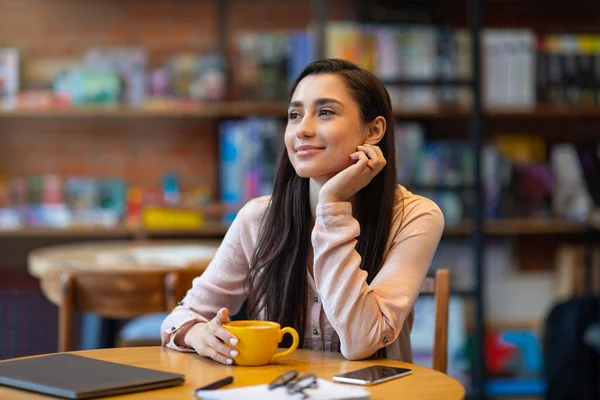 Мечтательная арабская леди с ноутбуком в кафе, пьет кофе и мечтает, учится или работает дистанционно — стоковое фото
