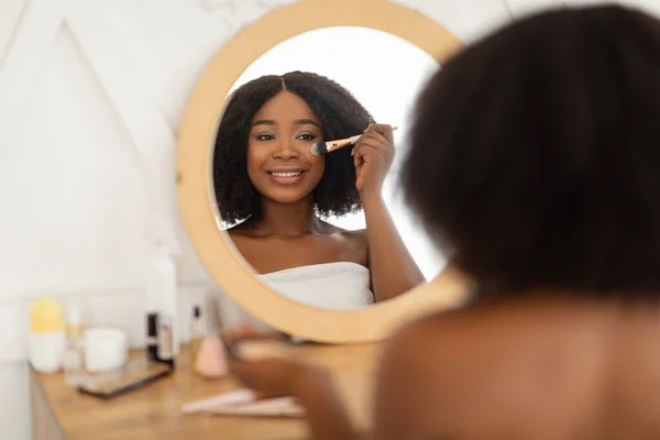 Attractive black lady applying decorative makeup in front of mirror, sitting at dressing table indoors — Stock Photo, Image