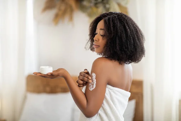 Trattamento di bellezza per una pelle sana. Splendida donna nera in asciugamano da bagno applicare crema per il corpo o burro sulla spalla a casa — Foto Stock