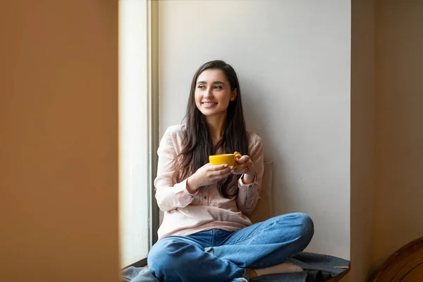 Ontspannen Arabische dame die 's ochtends koffie drinkt in het café, zit bij het raam op vensterbank, heeft pauze bij de studie, vrije ruimte — Stockfoto