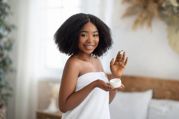 Zorgen voor de jonge huid. Sensuele duizendjarige zwarte dame aanbrengen gezichtscrème na douche, glimlachen op camera binnen — Stockfoto