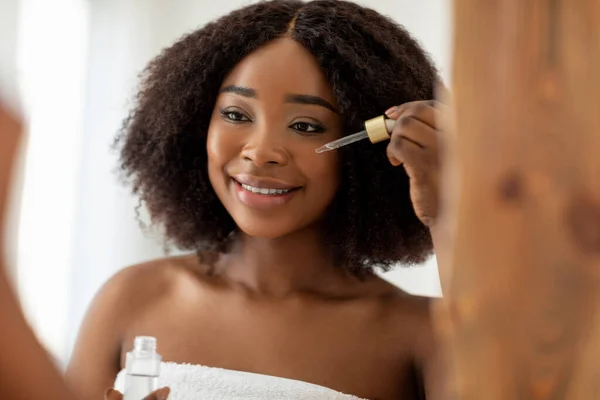 Alegre joven dama negra en toalla de baño usando suero facial cerca del espejo en casa — Foto de Stock