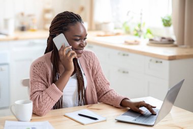 Mesafe çalışması. Mutfakta cep telefonu ve dizüstü bilgisayar kullanan neşeli siyah kadın.