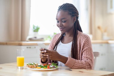 Mutlu Siyah Kadın Kahvaltı Yapıyor ve Akıllı Telefonu Mutfakta Kullanıyor