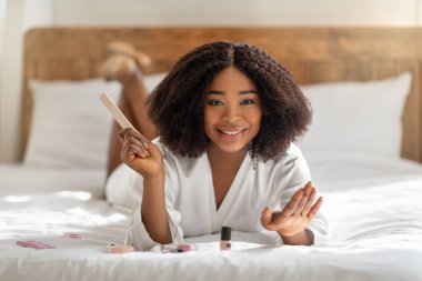 Milenyum siyahi bayanı manikür yapıyor, tırnak törpüsü tutuyor, kameraya gülümsüyor, evde yatakta yatıyor. Güzellik ve sağlık.