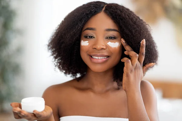Sensual young black lady applying moisturizing facial cream from jar near mirror at home. Beauty and natural cosmetics — Stock Photo, Image