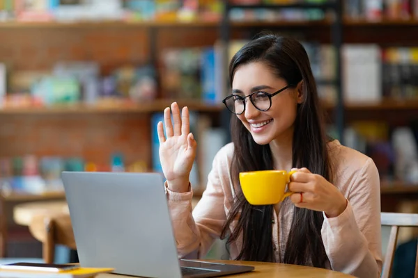 Amigável latina tendo videochamada do café, acenando com a mão para câmera do laptop e bebendo bebida quente — Fotografia de Stock