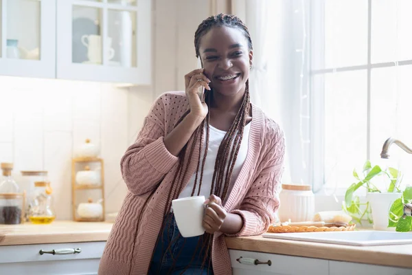 Domestic Leisure. African Lady Drinking Coffee And Talking On Cellphone At Home