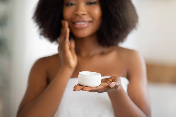 Conceito de tratamento de pele doméstico. Vista cortada da linda mulher negra segurando frasco de creme facial em casa, foco seletivo — Fotografia de Stock