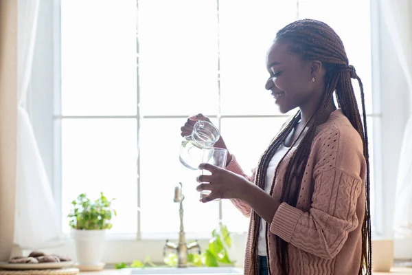 Allegro giovane signora nera che beve acqua in cucina a casa, tenendo brocca — Foto Stock