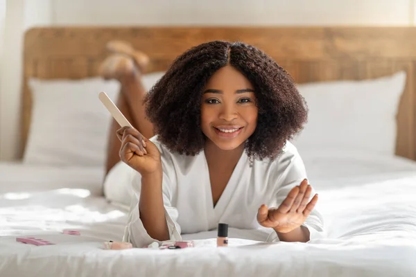 Millennial zwarte dame doet manicure, houdt nagelvijl vast, glimlacht naar de camera, ligt thuis op bed. Schoonheid en welzijn — Stockfoto