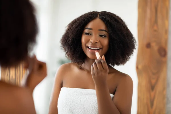 Retrato de belleza de una mujer negra bastante joven que aplica lápiz labial y mira el espejo en el interior. Productos de maquillaje ecológico — Foto de Stock