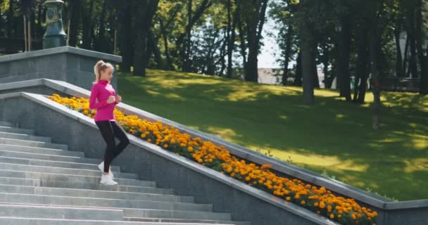Tysiącletnia blondynka w stroju sportowym biegnie po schodach miejskich rano, ćwiczy w parku publicznym, śledzi ujęcia — Wideo stockowe