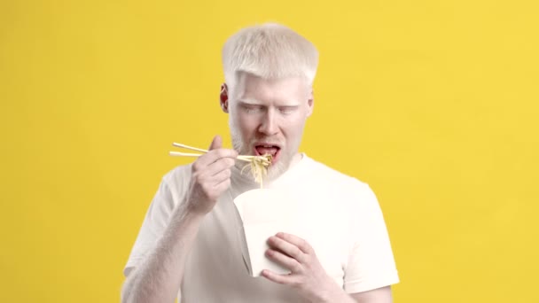 Парень-альбинос ест лапшу наслаждаясь нездоровой пищей на жёлтом фоне — стоковое видео