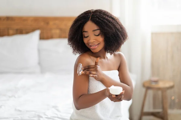 Belle dame noire enveloppée dans une serviette appliquer de la crème pour le corps sur son épaule après le bain dans la chambre — Photo