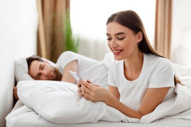 Cheating Wife Texting On Phone While Husband Sleeping In Bedroom clipart